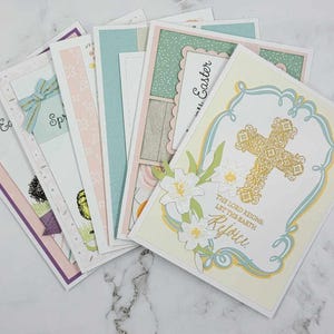 May include: A stack of handmade Easter cards with various designs, including a cross with flowers, a spring scene, and a simple Easter greeting. The cards are all white with pastel colors and gold accents.