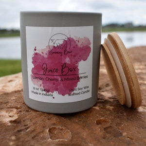 May include: A gray candle jar with a white label that says "Humming Bear Juice Box Lemon, Cherry, & Mixed Berries 12 oz (340g) Made in Indiana 100% Soy Wax Scented Candle". The candle jar has a wooden lid.