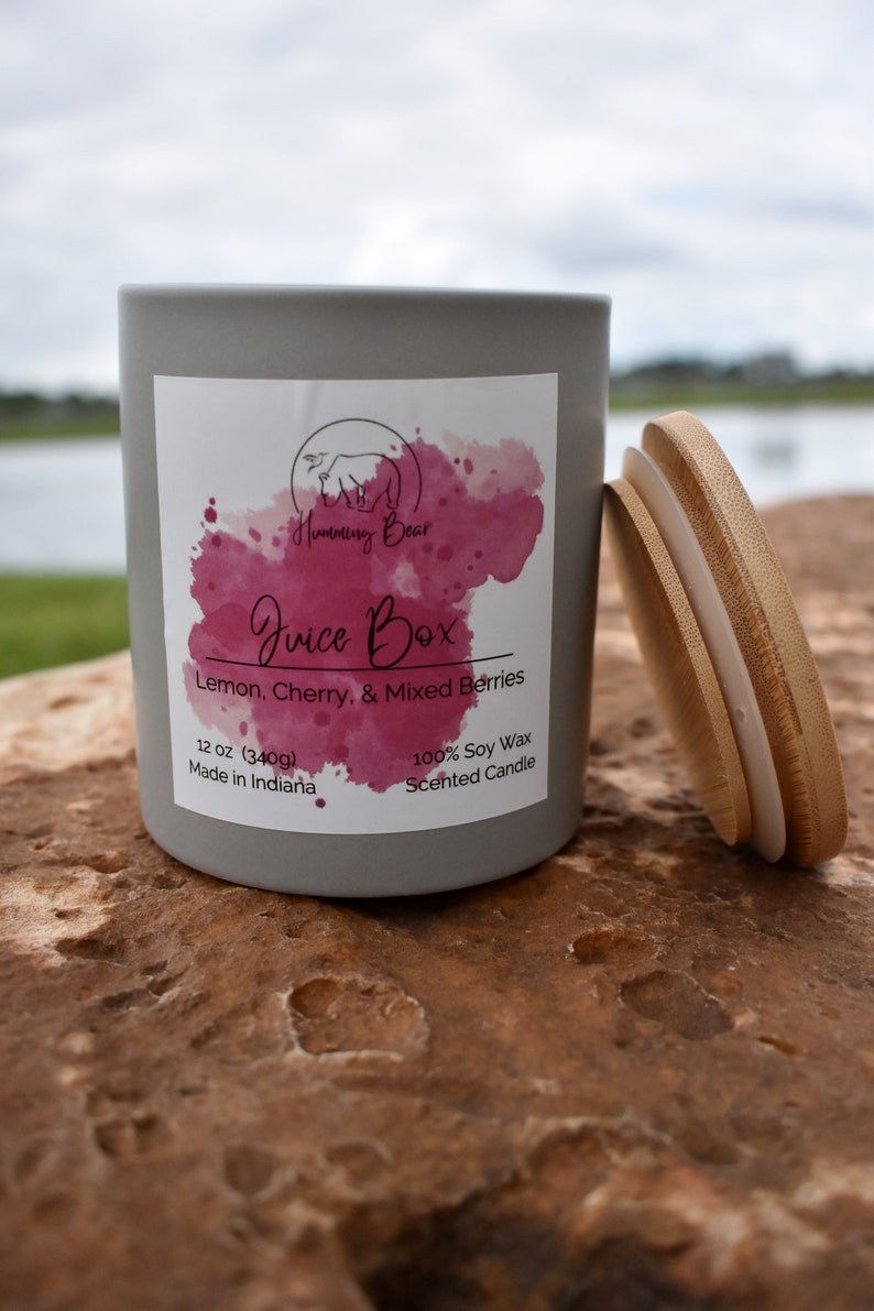 May include: A gray candle jar with a white label that says "Humming Bear Juice Box Lemon, Cherry, & Mixed Berries 12 oz (340g) Made in Indiana 100% Soy Wax Scented Candle". The candle jar has a wooden lid.