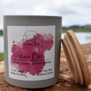May include: A gray candle jar with a white label that says "Humming Bear Juice Box Lemon, Cherry, & Mixed Berries 12 oz (340g) Made in Indiana 100% Soy Wax Scented Candle". The jar has a wooden lid.