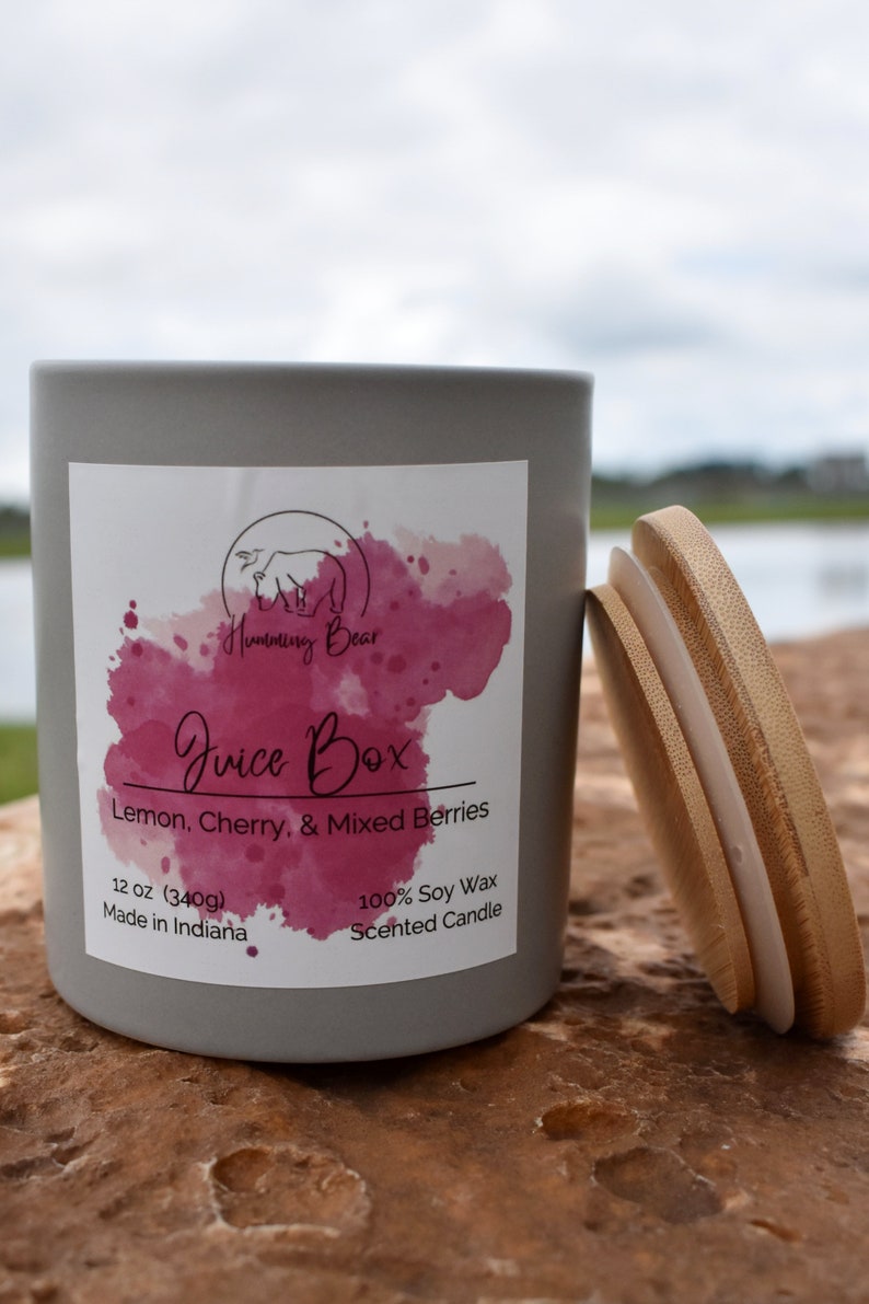 May include: A gray candle jar with a white label that says "Humming Bear Juice Box Lemon, Cherry, & Mixed Berries 12 oz (340g) Made in Indiana 100% Soy Wax Scented Candle". The jar has a wooden lid.