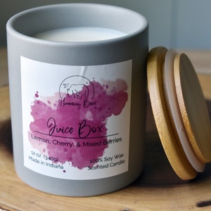May include: A gray ceramic candle jar with a white lid. The label says "Humming Bear Juice Box Lemon, Cherry, & Mixed Berries 12 oz (340g) Made in Indiana 100% Soy Wax Scented Candle".