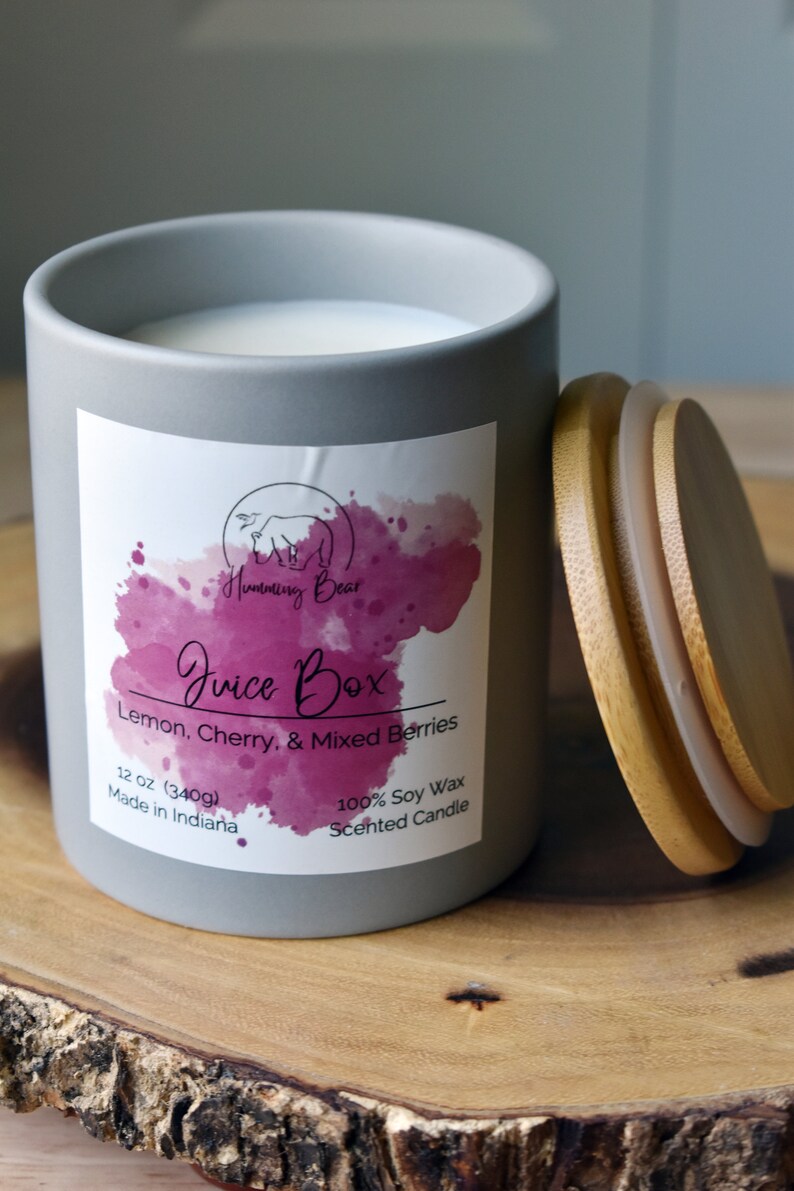 May include: A gray ceramic candle jar with a white lid. The label says "Humming Bear Juice Box Lemon, Cherry, & Mixed Berries 12 oz (340g) Made in Indiana 100% Soy Wax Scented Candle".