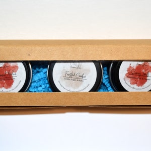 May include: A brown cardboard box with a window showing three small round tins of scented candles. The candles are labeled "Peppermint Mocha", "Frosted Cookie", and "Eucalyptus Mint".