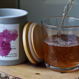 May include: A gray candle jar with a wooden lid. The label reads "Juice Box Lemon, Cherry, & Mixed Berries 12 oz (340g) Made in Indiana 100% Soy Wax Scented Candle". A glass of juice is being poured into a clear glass.