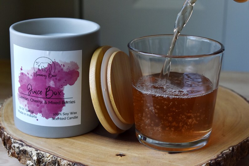 May include: A gray candle jar with a wooden lid. The label reads "Juice Box Lemon, Cherry, & Mixed Berries 12 oz (340g) Made in Indiana 100% Soy Wax Scented Candle". A glass of juice is being poured into a clear glass.