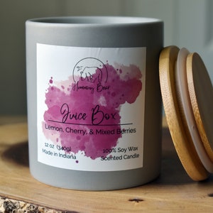 May include: A gray candle jar with a white label that says "Humming Bear Juice Box Lemon, Cherry, & Mixed Berries 12 oz (340g) Made in Indiana 100% Soy Wax Scented Candle". The candle jar has a wooden lid.