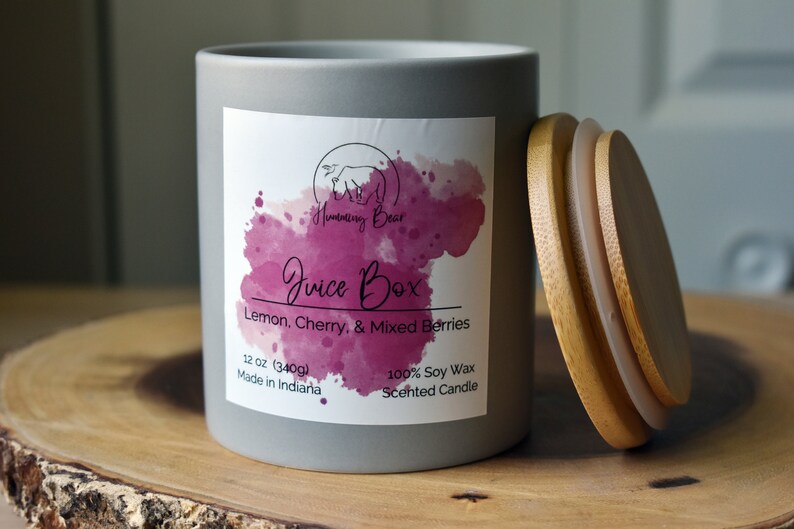 May include: A gray candle jar with a white label that says "Humming Bear Juice Box Lemon, Cherry, & Mixed Berries 12 oz (340g) Made in Indiana 100% Soy Wax Scented Candle". The candle jar has a wooden lid.