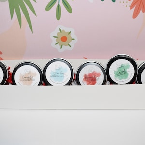 May include: Six small round metal tins with lids, each containing a candle. The tins are arranged in a row in a white box. The candle labels are visible and read "Sweet Rose", "Coconut Apricot", "Shine Them", "Rosemary & Mint", "Lilac", and "Twisted Cookie".