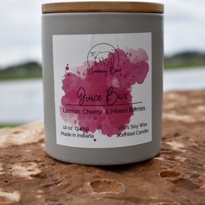 May include: A gray candle jar with a wooden lid. The label reads "Humming Bear Juice Box Lemon, Cherry, & Mixed Berries 12 oz (340g) Made in Indiana 100% Soy Wax Scented Candle".