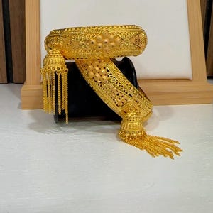 May include: Ornate gold-colored bangle bracelet with intricate filigree and tassel details. The bracelet is displayed on a black surface, showcasing its detailed design and craftsmanship. The jewelry piece is likely made of gold or a gold-toned metal.