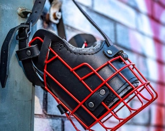May include: A black leather dog muzzle with a red metal cage. The muzzle is hanging on a hook.