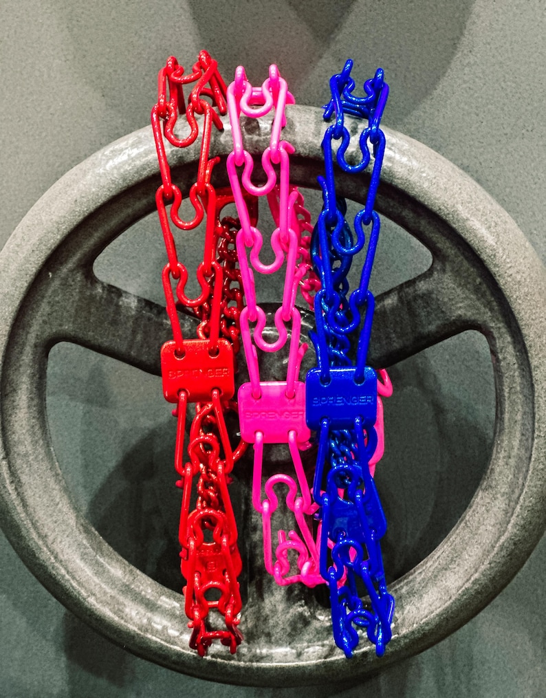 Op de afbeelding: Drie plastic kettingen in de kleuren rood, roze en blauw, elk met een zwart label met de tekst "SPRENGER". De kettingen hangen aan een metalen wiel.