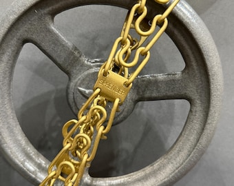 May include: A gold-colored chain with interlocking links, featuring a rectangular plate with the word "SPRENGER" engraved on it. The chain is set against a gray, circular background, creating a contrast in color and texture.
