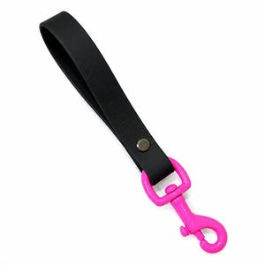 May include: A black wrist strap with a bright pink clasp. The strap is made of a textured, matte black material and is attached to a pink metal clasp with a swivel hook. A silver-colored rivet secures the strap.