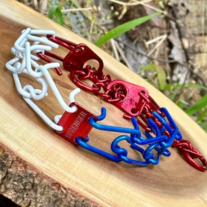 Puede incluir: Tres collares de adiestramiento canino en rojo, blanco y azul, dispuestos sobre una superficie de madera. Cada collar presenta un diseño de eslabones de cadena con una etiqueta metálica que dice "SPRENGER".