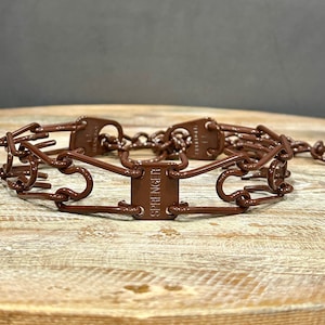 Puede incluir: Un collar de púas de metal marrón para perros, con el nombre de la marca "SPRENGER" impreso en la hebilla.
