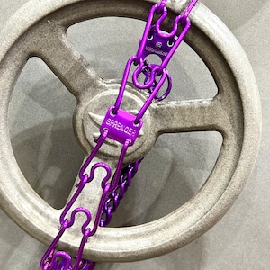 Puede incluir: Cadena de perro de metal morado con un gran anillo de metal redondo plateado. La cadena está unida a una rueda de metal plateada. La cadena tiene el texto "SPRENGER" en ella.