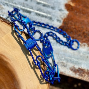 Puede incluir: Un collar de entrenamiento para perros de metal azul con la palabra "SPRENGER" en una placa rectangular. El collar está hecho de eslabones entrelazados y una cadena. El fondo es una mezcla de madera y una superficie de metal corrugado.