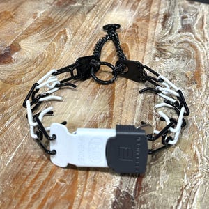 Puede incluir: Collar de púas negro y blanco con una cadena negra y una hebilla negra y blanca. La hebilla tiene el texto "SPRINGER".