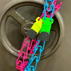 Puede incluir: Dos cadenas de seguridad de plástico verde y amarillo brillante con etiquetas negras "HS Germany Sprenger" unidas a una rueda de metal. Las cadenas están conectadas a una cadena de plástico rosa y azul.