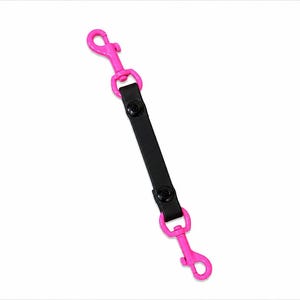 Puede incluir: Un divisor de correa para perros con cierres metálicos de color rosa brillante y una correa de cuero negro. La correa se asegura con dos broches negros. Los cierres están diseñados para sujetarse al collar o arnés de un perro.
