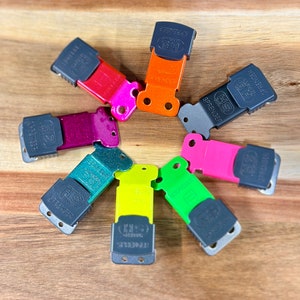 Op de afbeelding: Een verzameling kleurrijke Sprenger hondenriemclips, gerangschikt in een cirkelvormig patroon. De clips zijn in verschillende kleuren, waaronder rood, roze, oranje, grijs, groen en geel. Elke clip heeft de merknaam en het logo erop gedrukt.