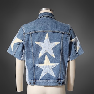 May include: A light blue denim jacket with short sleeves, adorned with large, cream-colored star appliqués. The back features two prominent stars, and smaller stars are on the sleeves. The jacket is displayed on a mannequin.