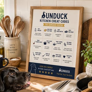 May include: A framed kitchen cheat sheet poster with a light wood frame, featuring the text SUNDUCK KITCHEN CHEAT CODES and THE SUNDUCK DOZEN, listing twelve cooking tips with icons, and the text GOOD DOG ENERGY and Float On. Trust The Duck.