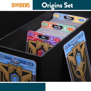 Pode incluir: Uma caixa preta para guardar cartas, com as etiquetas "DIVIDERS" e "Origins Set", contém vários divisores de cartas. Os divisores são codificados por cores com nomes como "Viktor" e "Lee Sin". As cartas têm um design dourado e azul com o texto "Origins Expansion Set #1."
