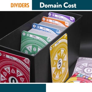 Pode incluir: Uma caixa preta cheia de cartas de jogo coloridas. As cartas são rotuladas com termos como "Fury", "Calm" e "Mind". A caixa tem o texto "DIVIDERS" e "Domain Cost" no topo. As cartas têm designs circulares.