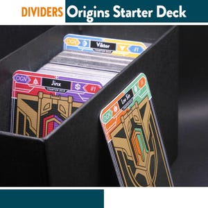 Pode incluir: Separadores de baralho de cartas pretas com o texto "DIVIDERS Origins Starter Deck". As cartas são rotuladas com nomes como "Viktor", "Jinx" e "Lee Sin". As cartas têm detalhes dourados, vermelhos, roxos e laranja. As cartas estão dentro de uma caixa preta.