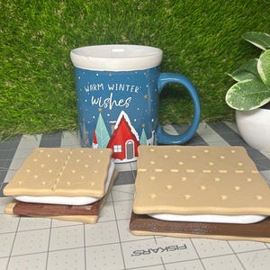 May include: Two graham cracker s'mores with chocolate and marshmallow filling. The s'mores are on a blue and gray cutting mat. A blue mug with the text "Warm Winter Wishes" is in the background.