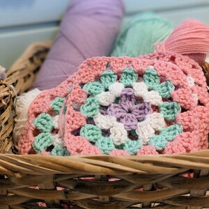 May include: A wicker basket filled with colorful yarn and crocheted squares in pink, green, white, and purple. The yarn is in various shades of pink, green, and white.