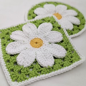 Könnte beinhalten: Zwei handgefertigte gehäkelte Untersetzer mit Gänseblümchen-Motiv. Einer ist quadratisch, der andere rund. Beide zeigen eine weiße Gänseblümchen mit gelber Mitte auf grünem Hintergrund, weiß umrandet. Ideal für die Heimdekoration.