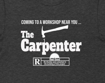 May include: A dark gray t-shirt with a white graphic design that says "Coming to a workshop near you... The Carpenter" with a hammer and saw blade graphic. The graphic also includes a rating label that says "R Real Good My thumbs were smashed in the building of this project."