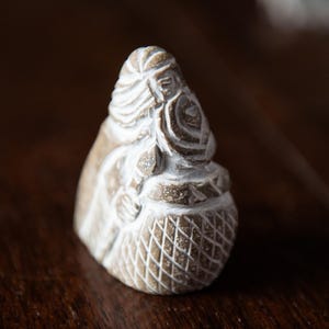 Puede incluir: Una figura de piedra tallada de una persona con la cabeza cubierta y sosteniendo una cesta. La figura es de color marrón claro con detalles blancos, y la cesta tiene un patrón de diamantes. La figura está sobre una superficie de madera oscura.