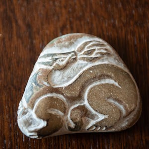 Könnte beinhalten: Ein geschnitzter Stein mit einem stilisierten Tierdesign. Der Stein ist eine Mischung aus Braun, Beige und Weiß, wobei die geschnitzten Details weiß hervorgehoben sind. Das Kunstwerk zeigt ein liegendes Tier, möglicherweise eine Katze oder ein ähnliches Wesen.
