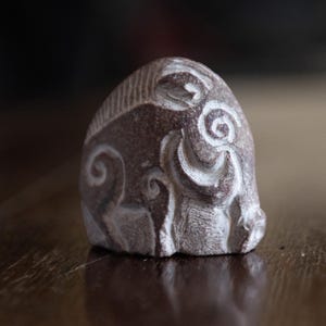 Könnte beinhalten: Eine kleine, handgeschnitzte Steinskulptur eines Widders mit komplizierten, wirbelnden Mustern. Die Skulptur ist aus dunkelbraunem Stein mit weißen Details gefertigt.