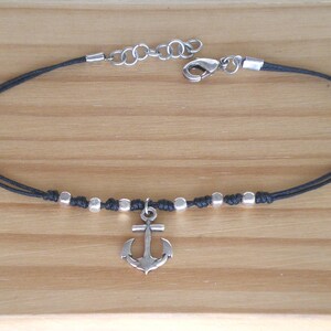 May include: An anklet with a black cord and silver-colored beads and an anchor charm. The anklet has a lobster clasp closure and is displayed on a wooden surface.
