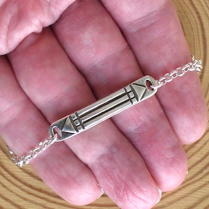 Op de afbeelding: Een zilveren armband met een rechthoekige hanger met geometrische ontwerpen. De hanger is verbonden met een delicate ketting. De armband wordt in een hand gehouden, waarbij de details en het vakmanschap van de sieraden worden getoond.