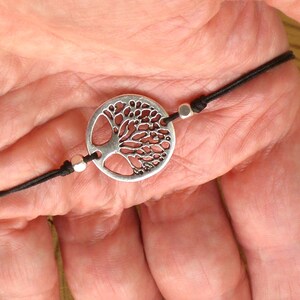 Puede incluir: Un brazalete de cordón negro con un dije de plata en forma de árbol de la vida. El dije es redondo y presenta un diseño detallado de un árbol con ramas y hojas.