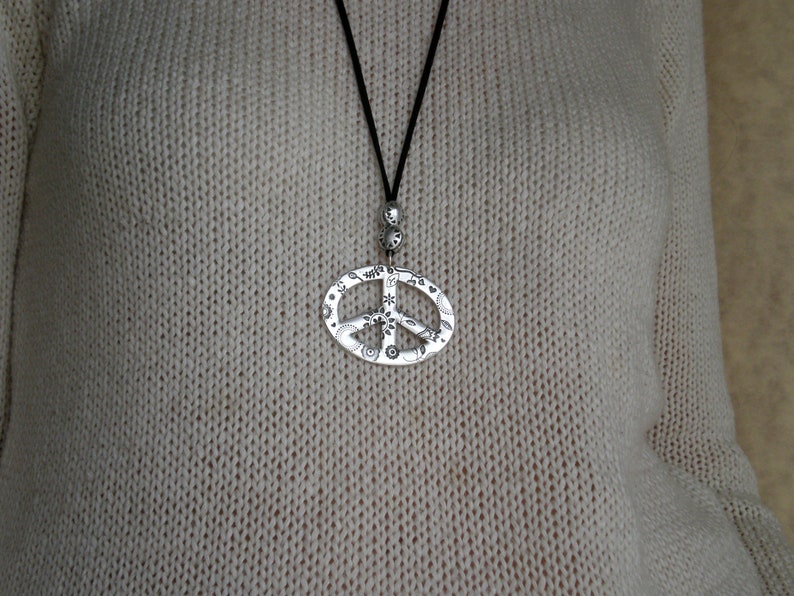 May include: A silver peace sign pendant necklace with a black cord. The pendant is detailed with small symbols and designs.