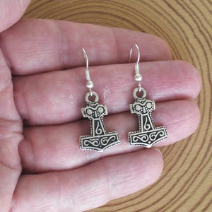 Pode incluir: Um par de brincos de metal prateado com um pendente em forma de martelo de Mjolnir. Cada pendente apresenta padrões de nós em relevo intrincados e um detalhe circular central, suspenso por um gancho de metal prateado simples.