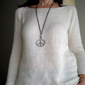 May include: A white knitted sweater with a long sleeve and a scoop neck. The sweater has a silver peace sign pendant on a black cord.