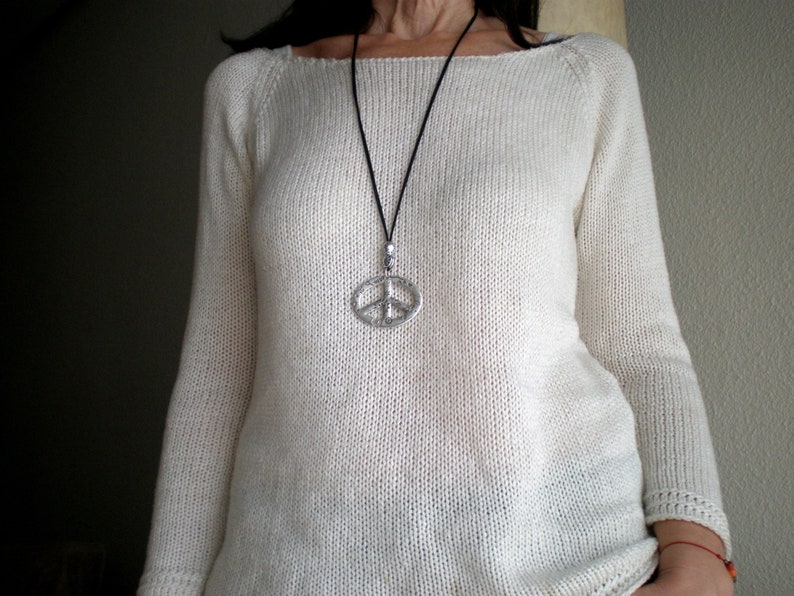 May include: A white knitted sweater with a long sleeve and a scoop neck. The sweater has a silver peace sign pendant on a black cord.