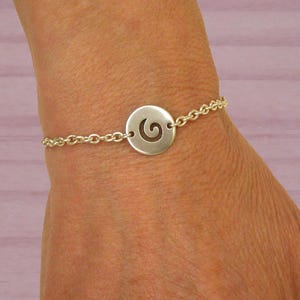 Puede incluir: Pulsera de plata con un colgante circular con un diseño en espiral. El colgante está conectado a una fina pulsera de cadena. La pulsera se muestra en una muñeca. Una joya sencilla y elegante. Fondo rosa claro.