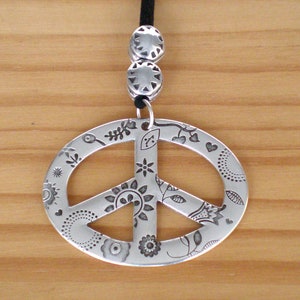 May include: A silver peace sign pendant necklace with a black cord. The peace sign is decorated with a floral design.