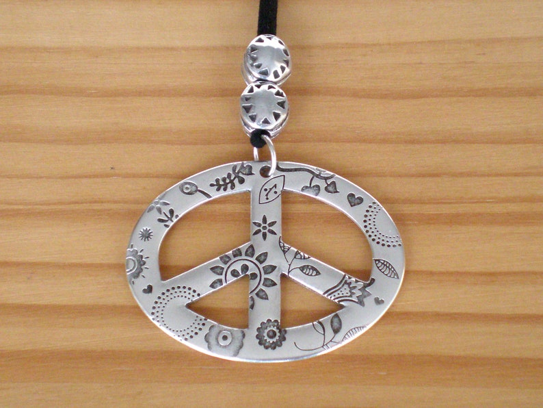May include: A silver peace sign pendant necklace with a black cord. The peace sign is decorated with a floral design.