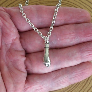Könnte beinhalten: Eine silberfarbene Halskette mit einem fußförmigen Anhänger. Der Anhänger ist an einer Kette befestigt. Die Halskette wird in der Hand einer Person gehalten.